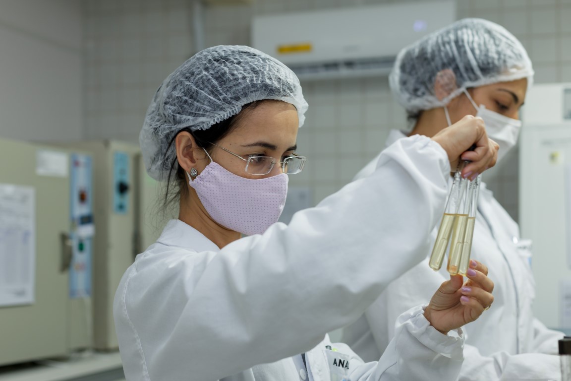 Analistas da GTA com touca e máscara examinando tubos de ensaio no laboratório de microbiologia